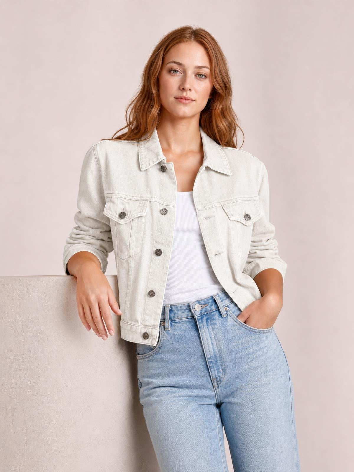 A model leans against a ledge wearing an off-white button-up long sleeve denim jacket with a collared front, chest flap pockets, and relaxed silhouette.