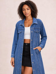 A model poses in a blue longline denim jacket with long sleeves, a collared button front, chest flap pockets, and side pockets.
