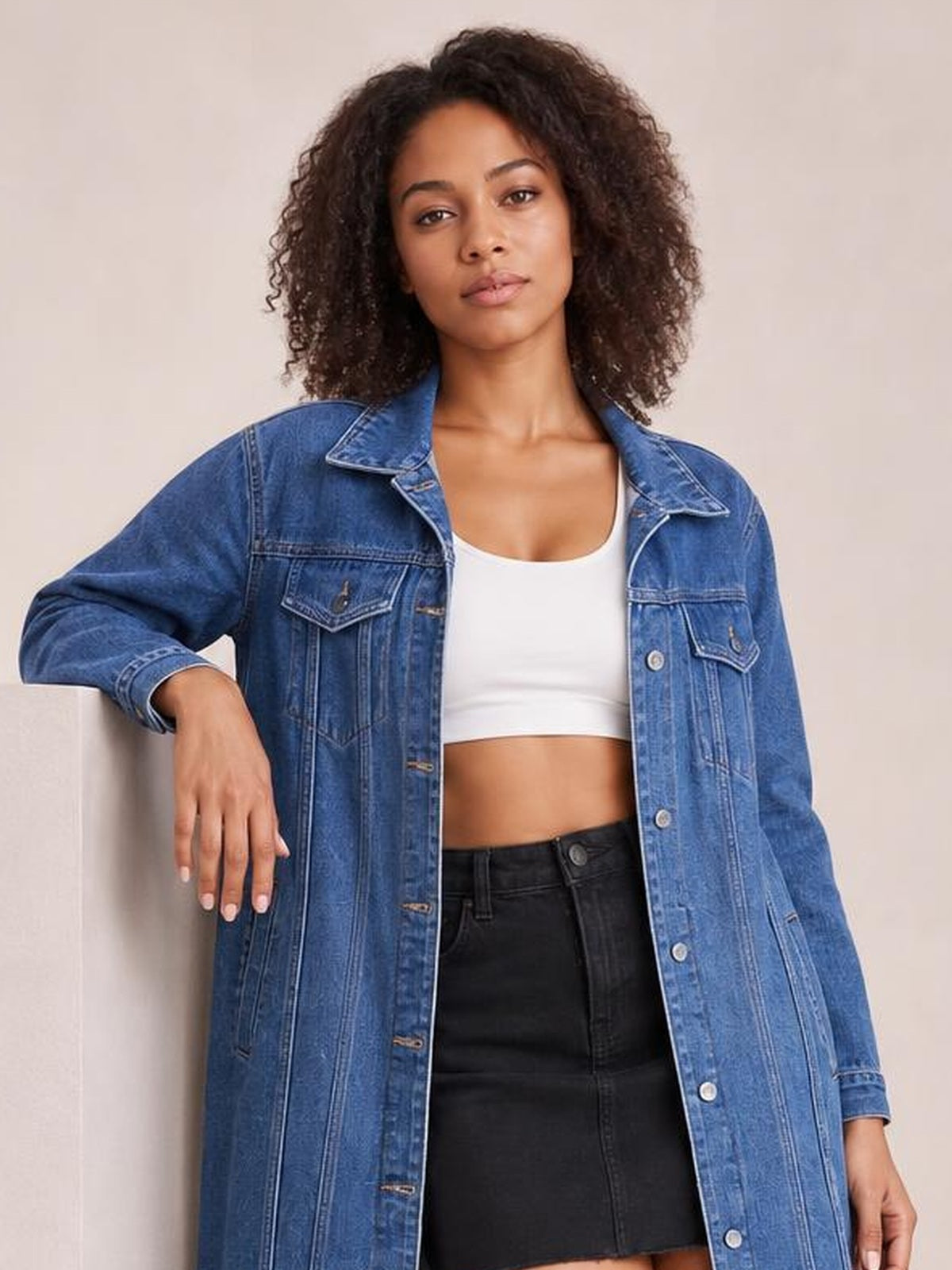 Model wearing a blue button-up longline denim jacket with long sleeves, flap pockets, pointed collar, vertical seam detailing, and a relaxed silhouette.