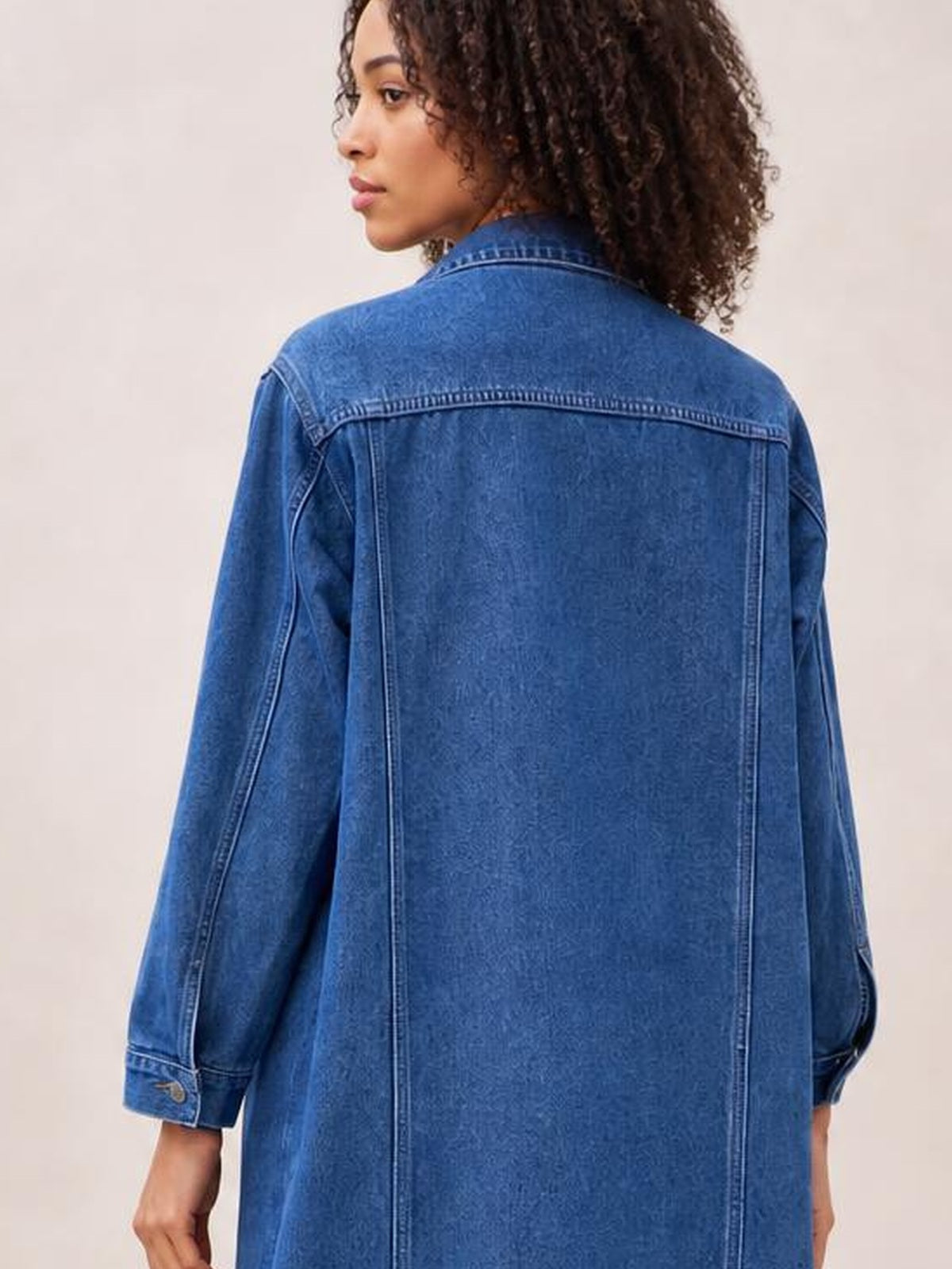Model shown from behind wearing a blue longline denim jacket with long sleeves, a pointed collar, relaxed silhouette, and visible seam detailing.