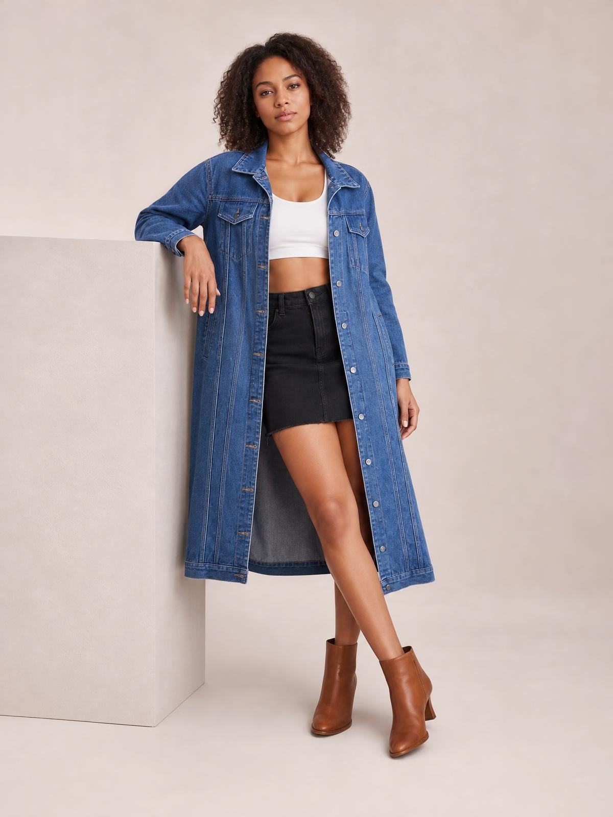 Model wearing a blue longline denim jacket with long sleeves, button closure, chest pockets, and a relaxed straight silhouette while leaning with one arm on a ledge.