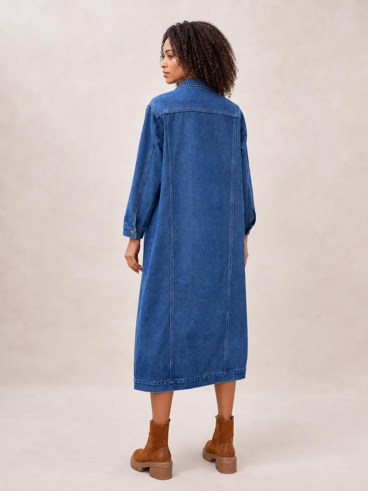 Model shown from the back in a blue longline denim jacket with long sleeves, button front, seamed construction, and a straight mid-calf silhouette.