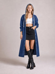 A model stands in a blue denim longline hooded shirt with button front, long sleeves, relaxed silhouette, and one hand tucked into a pocket.