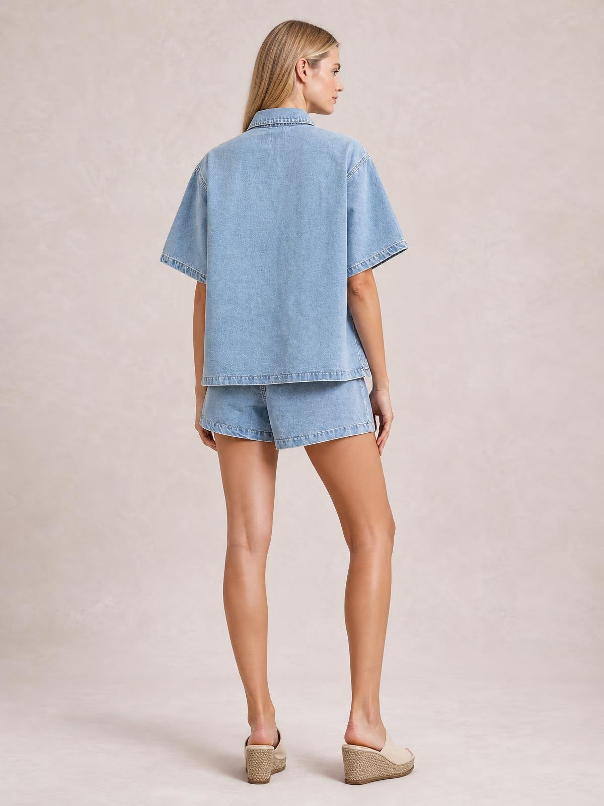 Model shown from the back in a light blue denim set with a loose button-up short-sleeve top and matching pocketed shorts, standing in a relaxed pose.