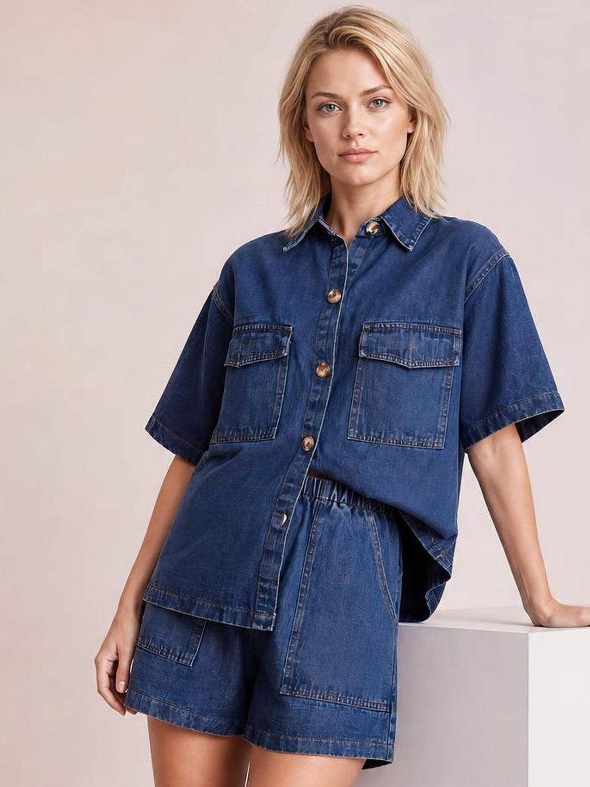 A model poses in a dark blue denim set with a button-up short-sleeve shirt, collared design, patch pockets, and matching pocketed shorts.