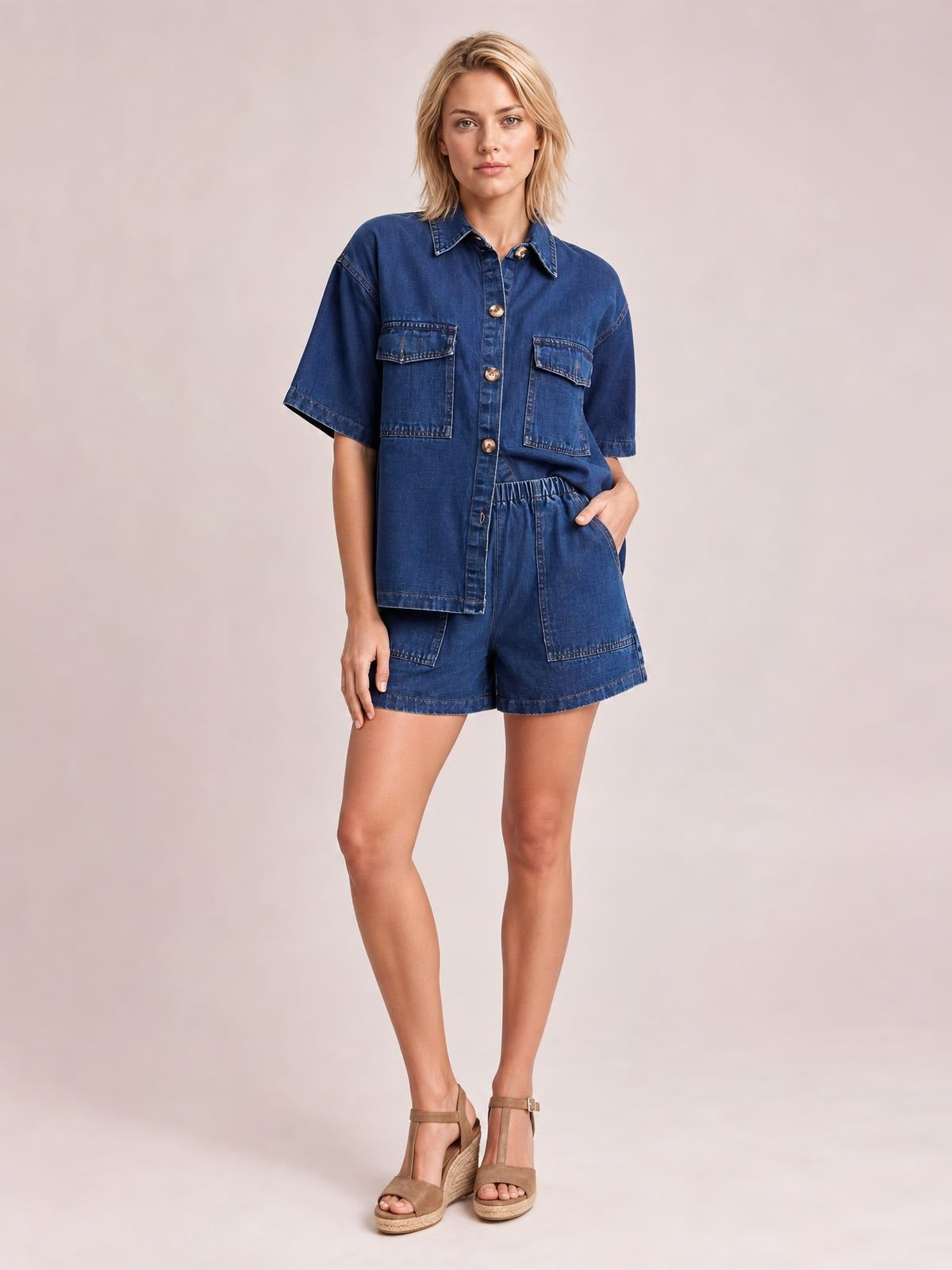 A model poses in a dark blue denim set with a button-up short-sleeve shirt, flap chest pockets, and relaxed pocketed shorts with an elastic waist.