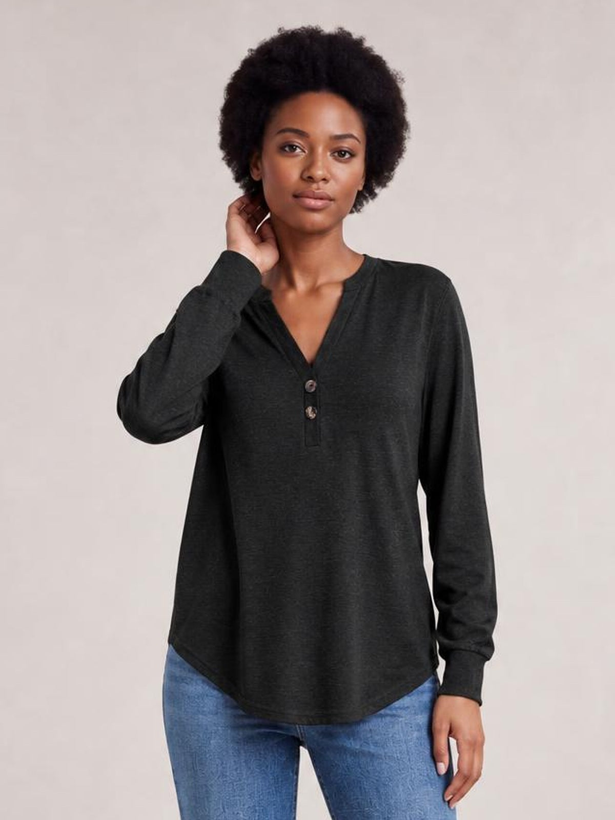 Model wearing a charcoal buttoned notched-neck long-sleeve top with a relaxed silhouette, curved hem, and ribbed cuffs, posing with one hand near her neck.