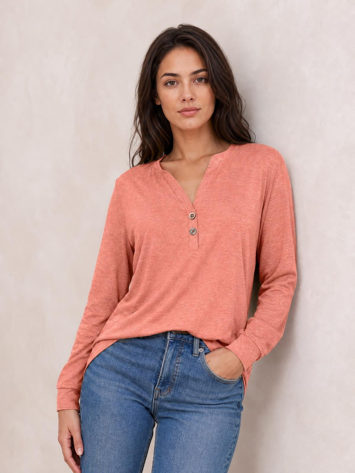 Model wears a coral buttoned notched-neck long-sleeve top with a relaxed silhouette, long cuffs, and a short front placket, posing with one hand in her jeans pocket.