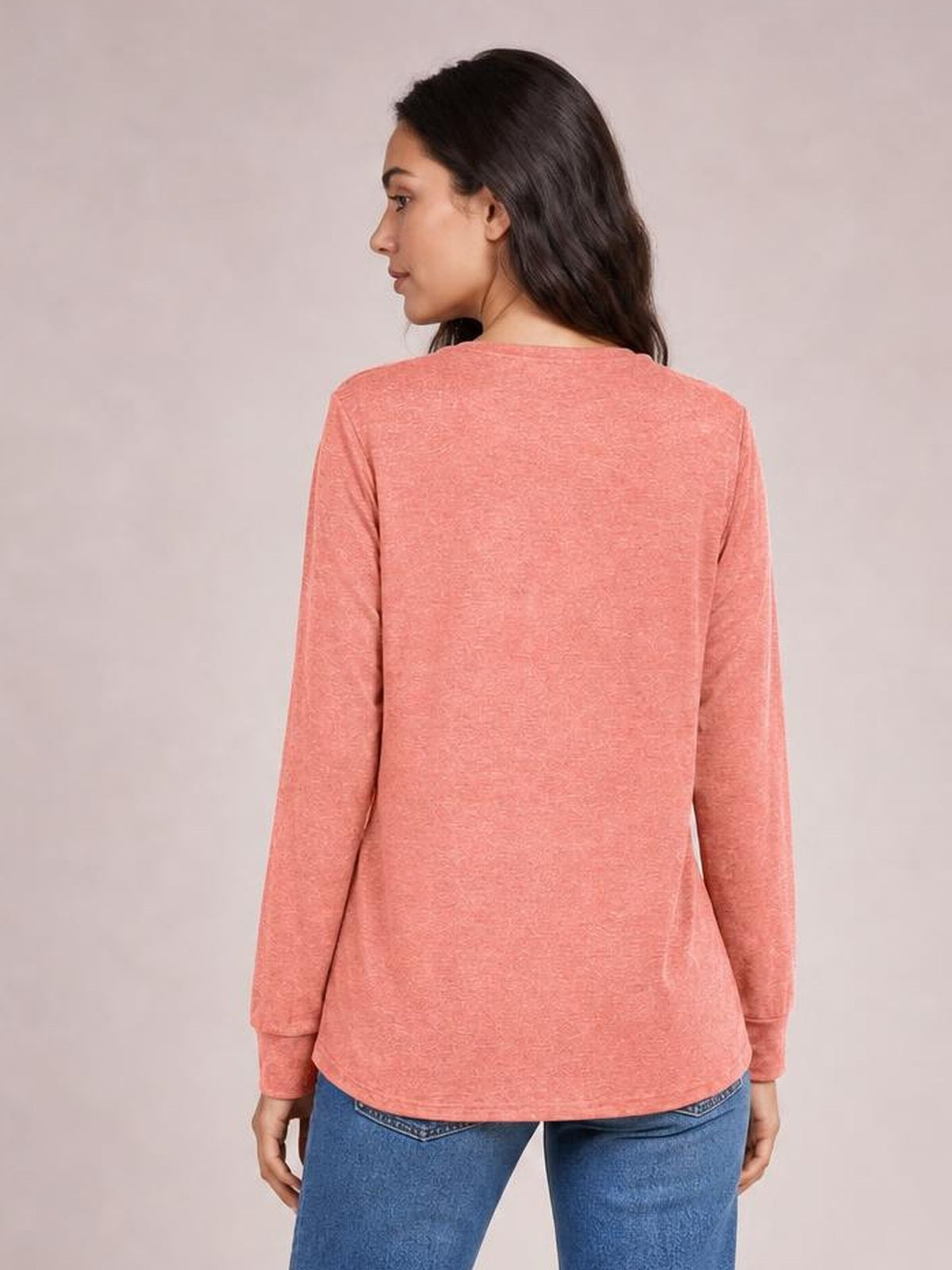 A model shows the back of a coral pink long-sleeve top with a relaxed, straight silhouette, standing with her arms at her sides.
