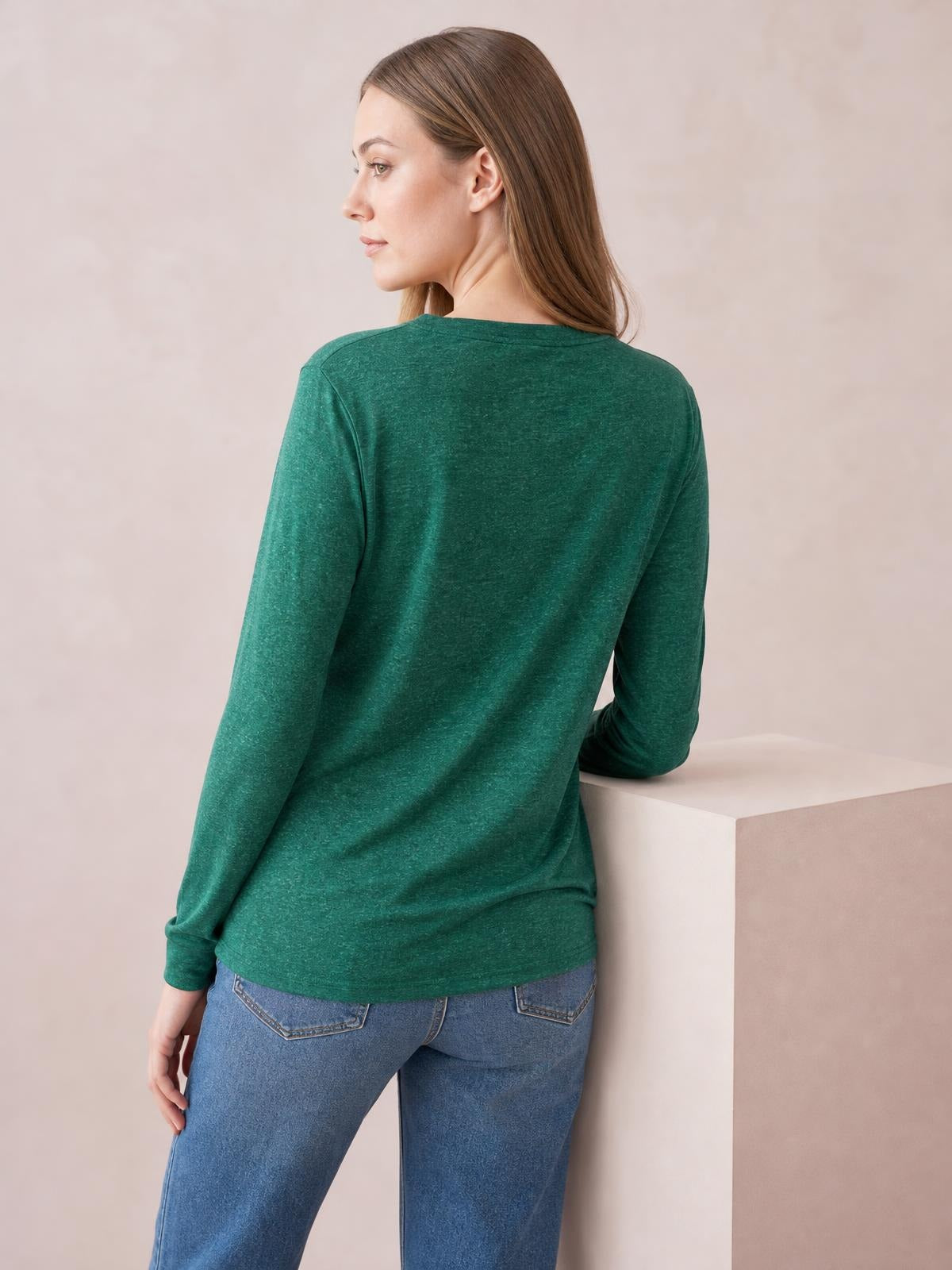 Model turned away wears a deep green long sleeve top with a relaxed straight silhouette and hip-length hem, resting one arm on a ledge.