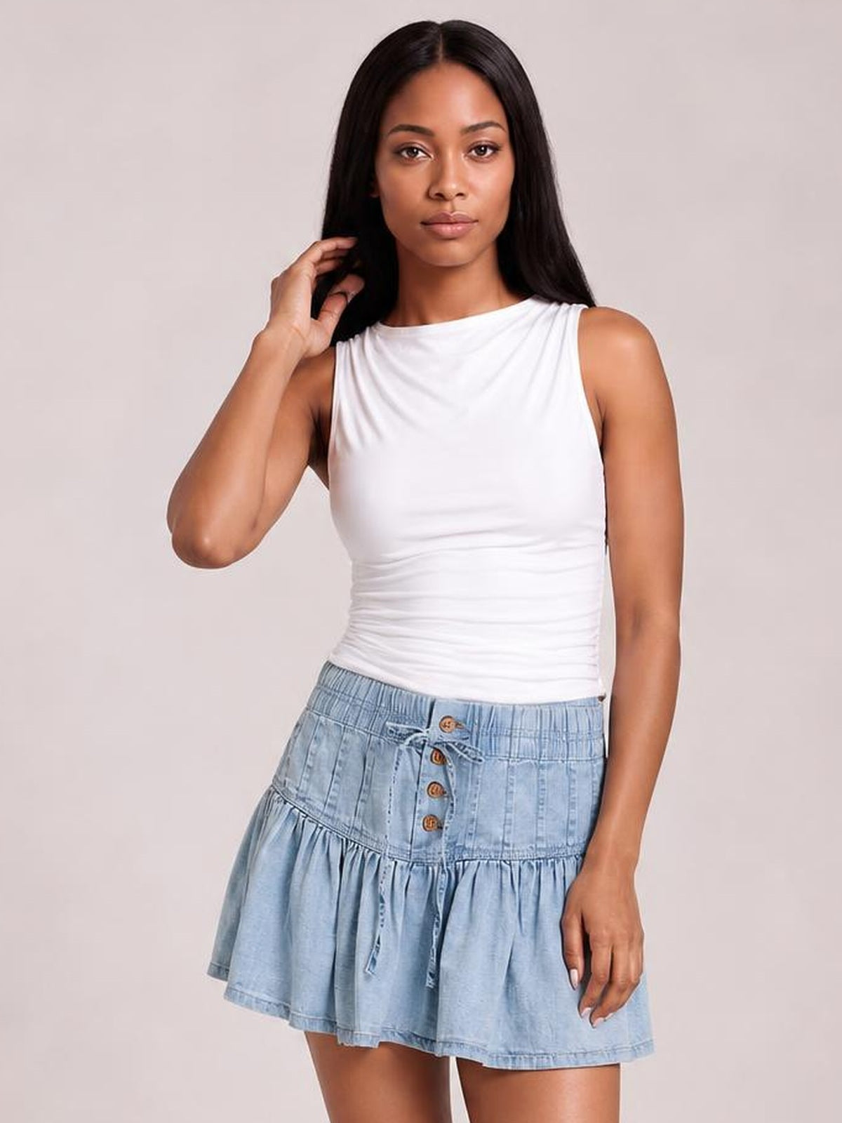 Model poses with one hand in her hair wearing a light blue buttoned ruffled mini denim skirt with a drawstring waist and tiered flared silhouette.