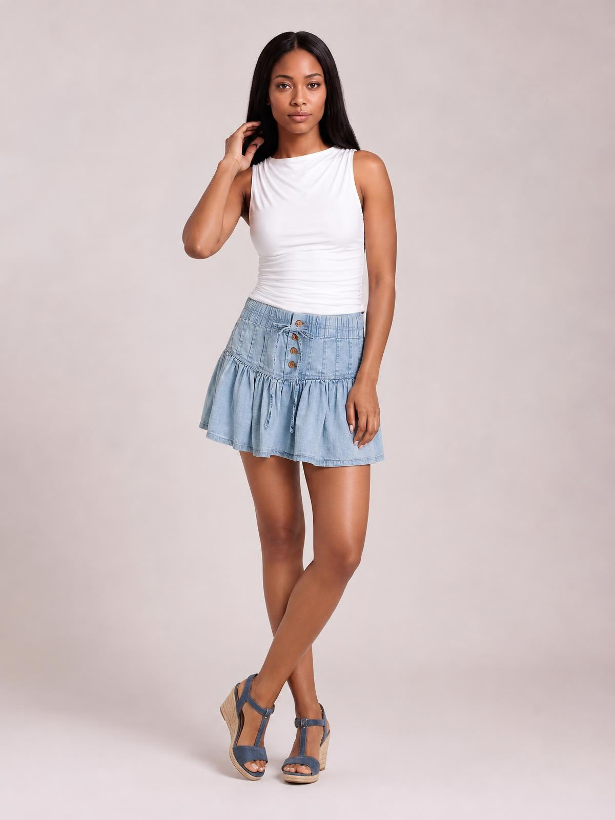 Model poses with crossed legs in a light blue buttoned ruffled mini denim skirt featuring a gathered waistband, front buttons, and drawstring ties.