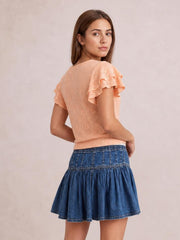 A model looks over her shoulder wearing a blue denim mini skirt with a gathered ruffled tier, paneled waistband, and flared silhouette.