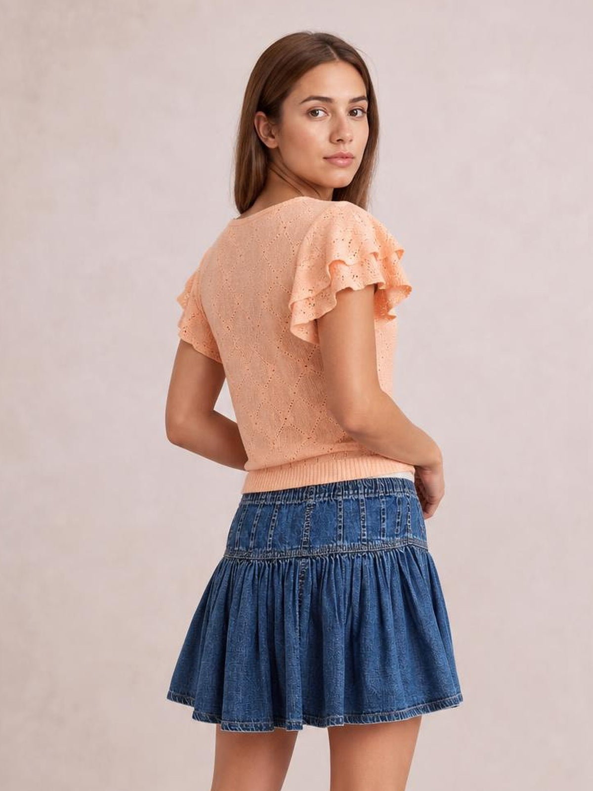 A model looks over her shoulder wearing a blue denim mini skirt with a gathered ruffled tier, paneled waistband, and flared silhouette.