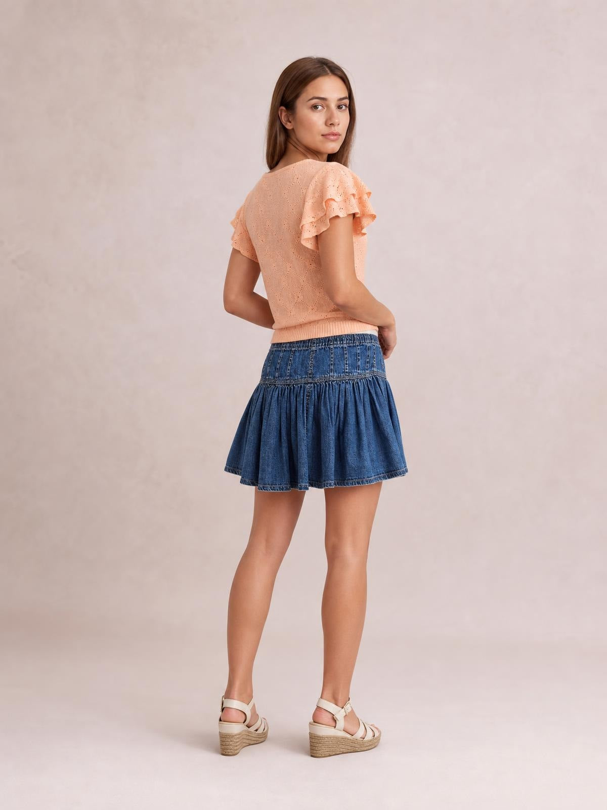 A model looks over her shoulder in a blue denim mini skirt with a gathered ruffled tier, high waistband, and flared silhouette.