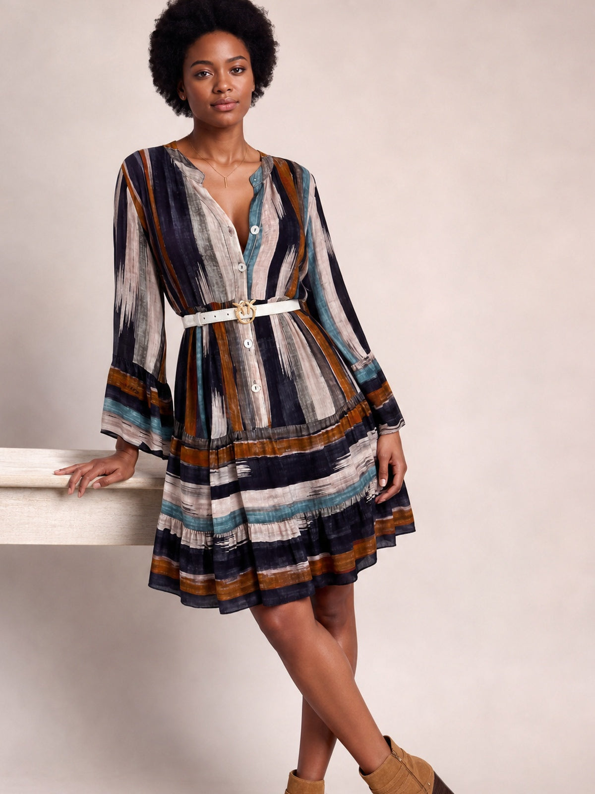 Model poses with crossed legs in a button-front striped long-sleeve mini dress featuring navy, rust, teal, and cream tones, a defined waist, and softly flared skirt.