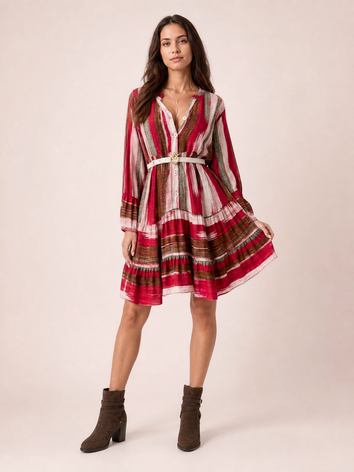 Model wearing a red, brown, and gray striped button-front long-sleeve mini dress with a belted waist and softly flared skirt, holding the hem to show its movement.