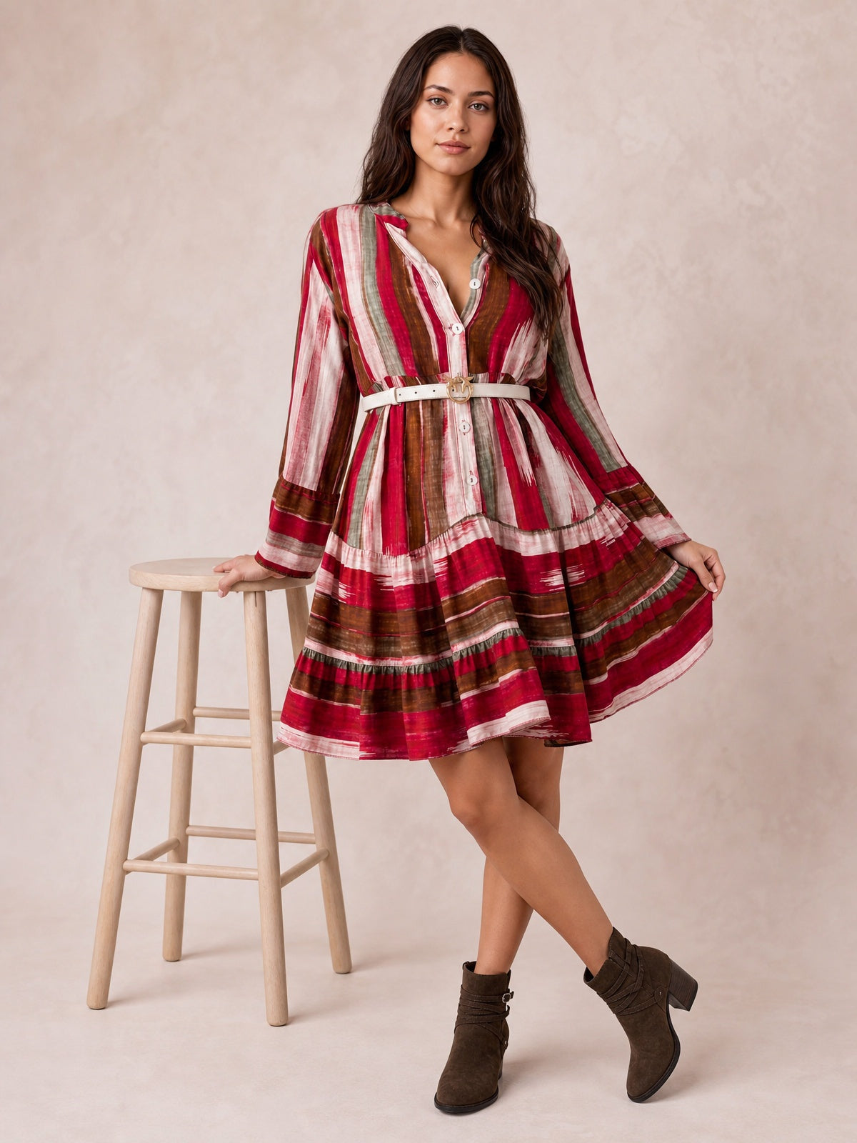 A model poses with one hand on a stool in a multicolored striped mini dress featuring long sleeves, a button front, belted waist, and softly flared skirt.