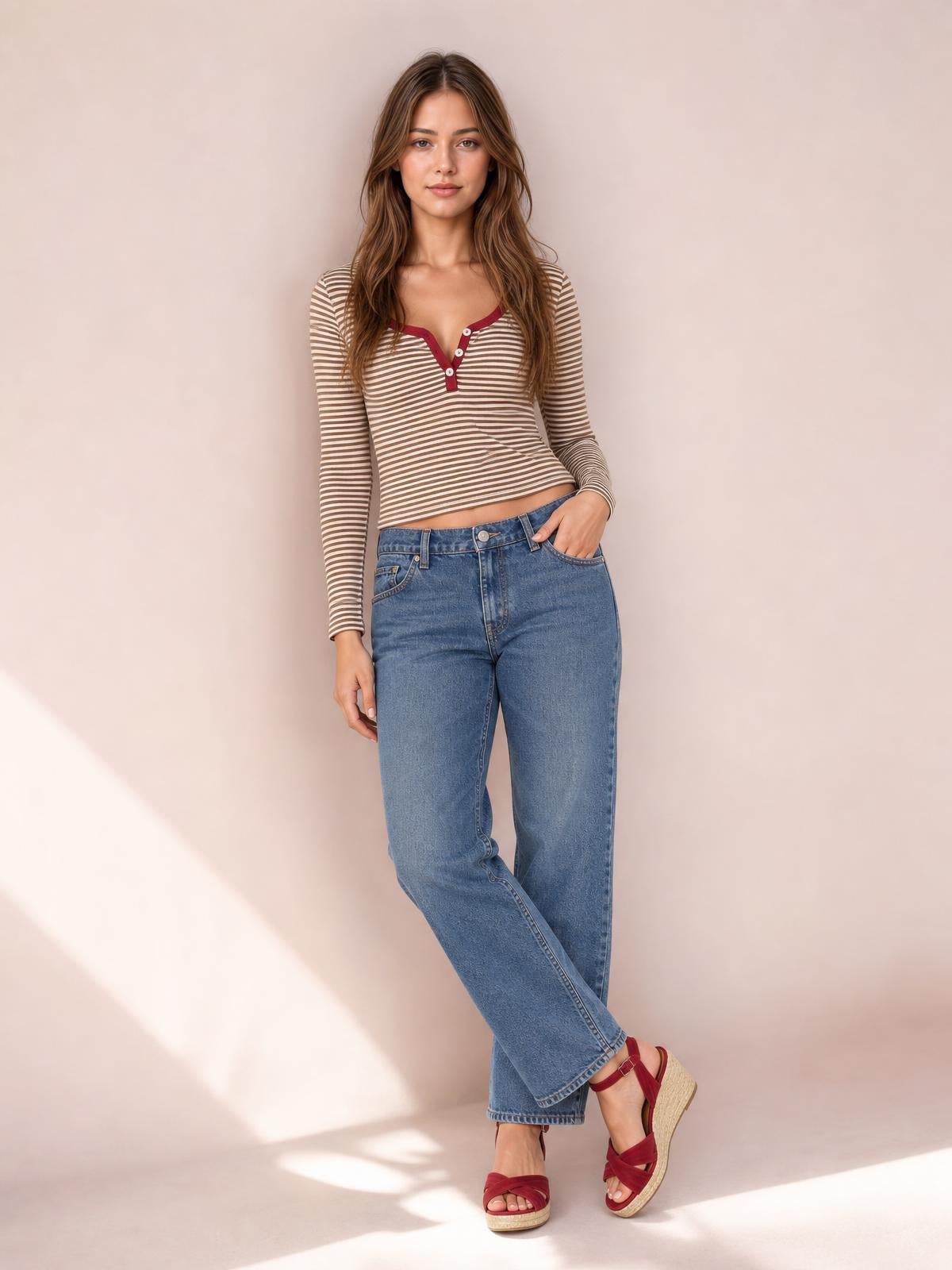 A model poses with crossed legs in a fitted beige-and-white striped long sleeve T-shirt featuring a buttoned Henley neckline and burgundy trim.