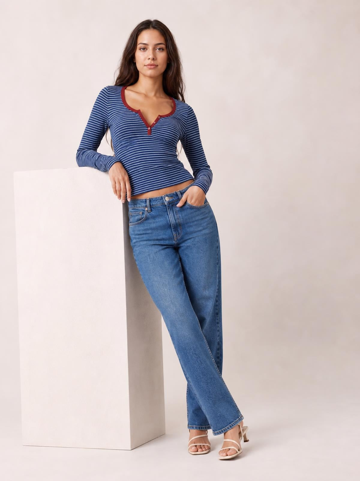 Model leans against a pedestal wearing a fitted navy-and-white striped long sleeve T-shirt with a buttoned notched neckline and red trim.