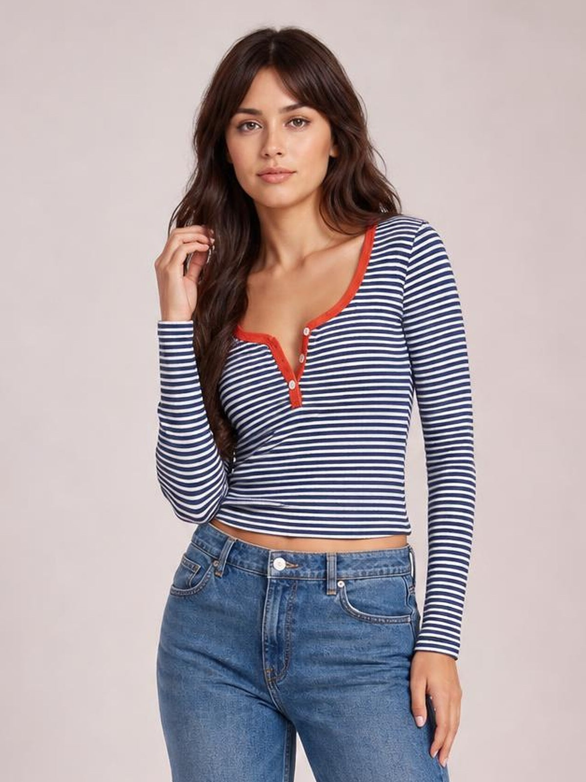 A model poses in a fitted navy-and-white striped long sleeve T-shirt with red-trimmed buttoned neckline and cropped silhouette, touching her hair.
