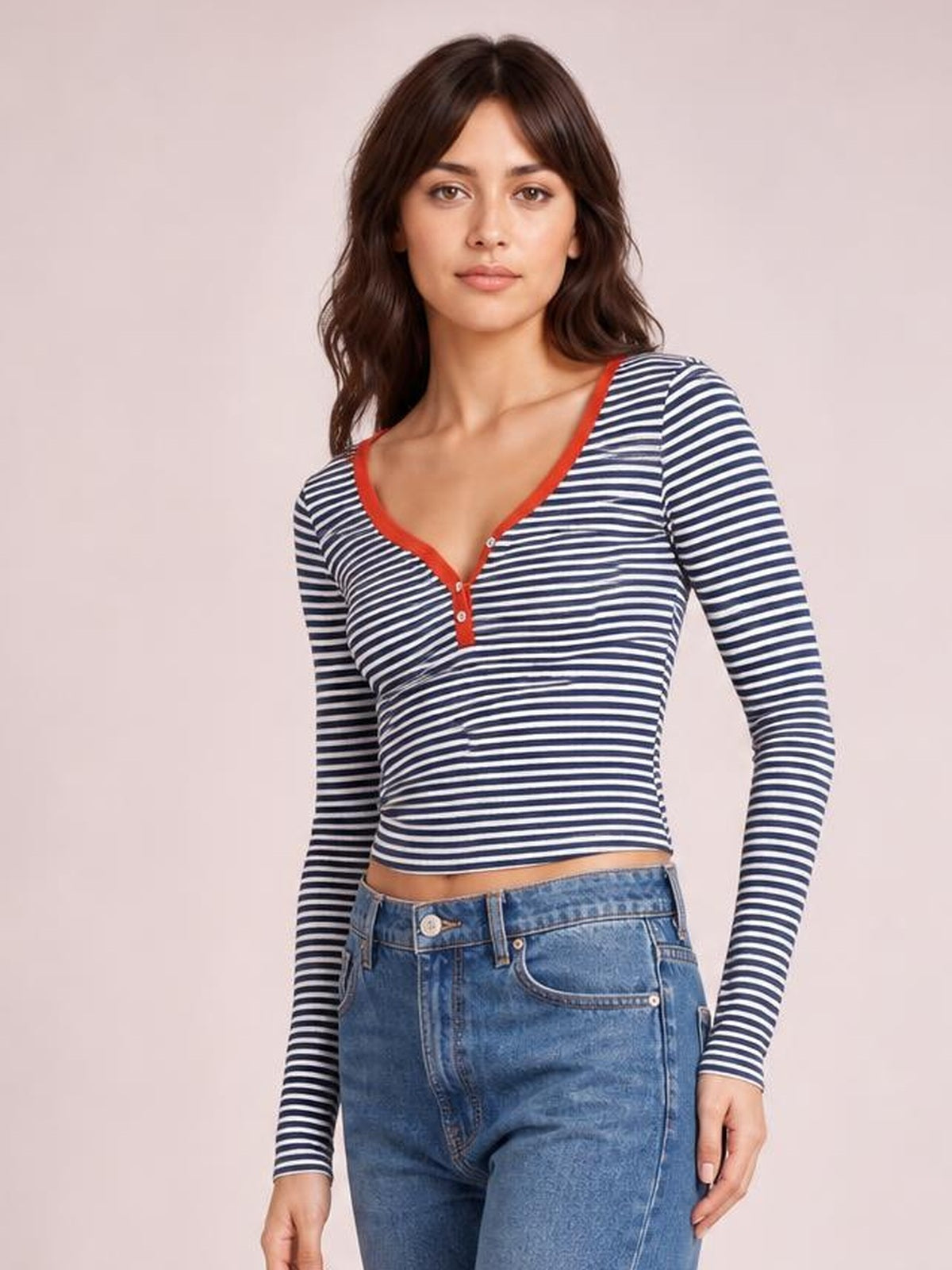 Model wearing a navy and white striped long sleeve T-shirt with a red-trimmed buttoned neckline and fitted cropped silhouette, standing with one hand at her side.