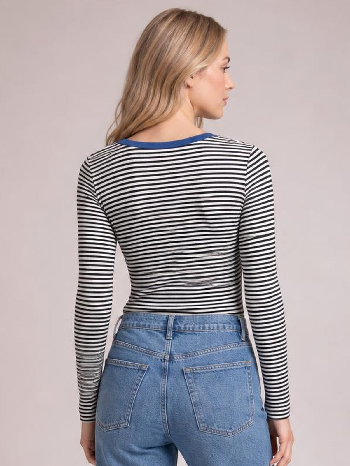 A model stands in side profile wearing a fitted black-and-white striped long sleeve T-shirt with a blue-trimmed round neckline and relaxed arms.