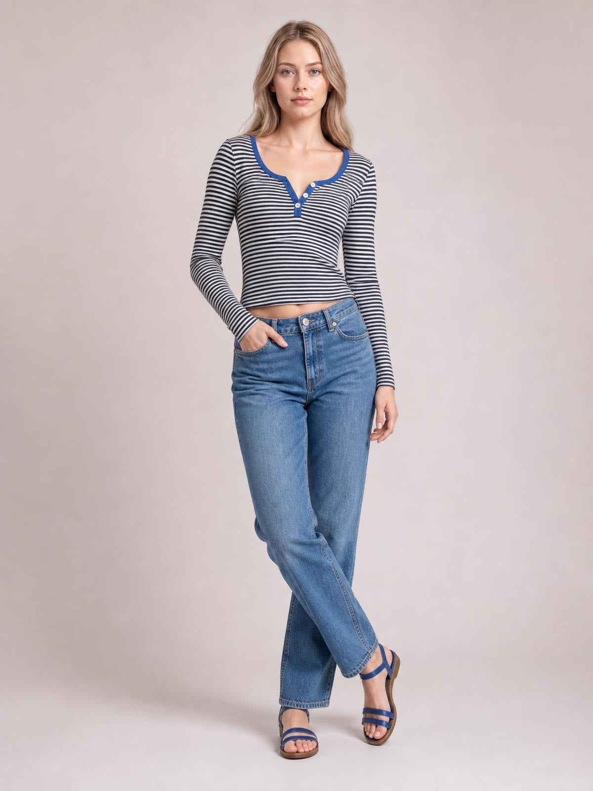 Model wearing a fitted black-and-white striped long sleeve T-shirt with a buttoned blue-trimmed neckline, posing with one hand in her jeans pocket.