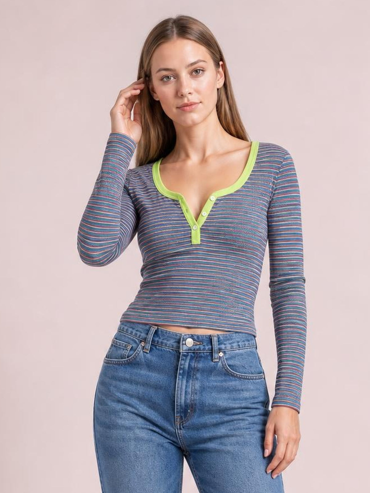 A model poses with one hand near her hair, wearing a fitted striped long sleeve T-shirt in blue, pink, and gray with a green button placket.