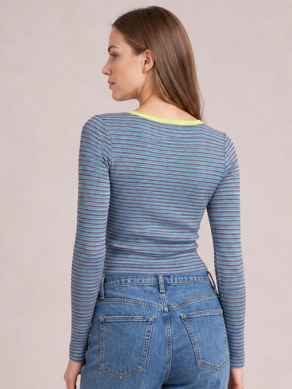 A model looks over her shoulder wearing a fitted striped long sleeve T-shirt with blue, teal, and purple horizontal stripes and a rounded neckline.