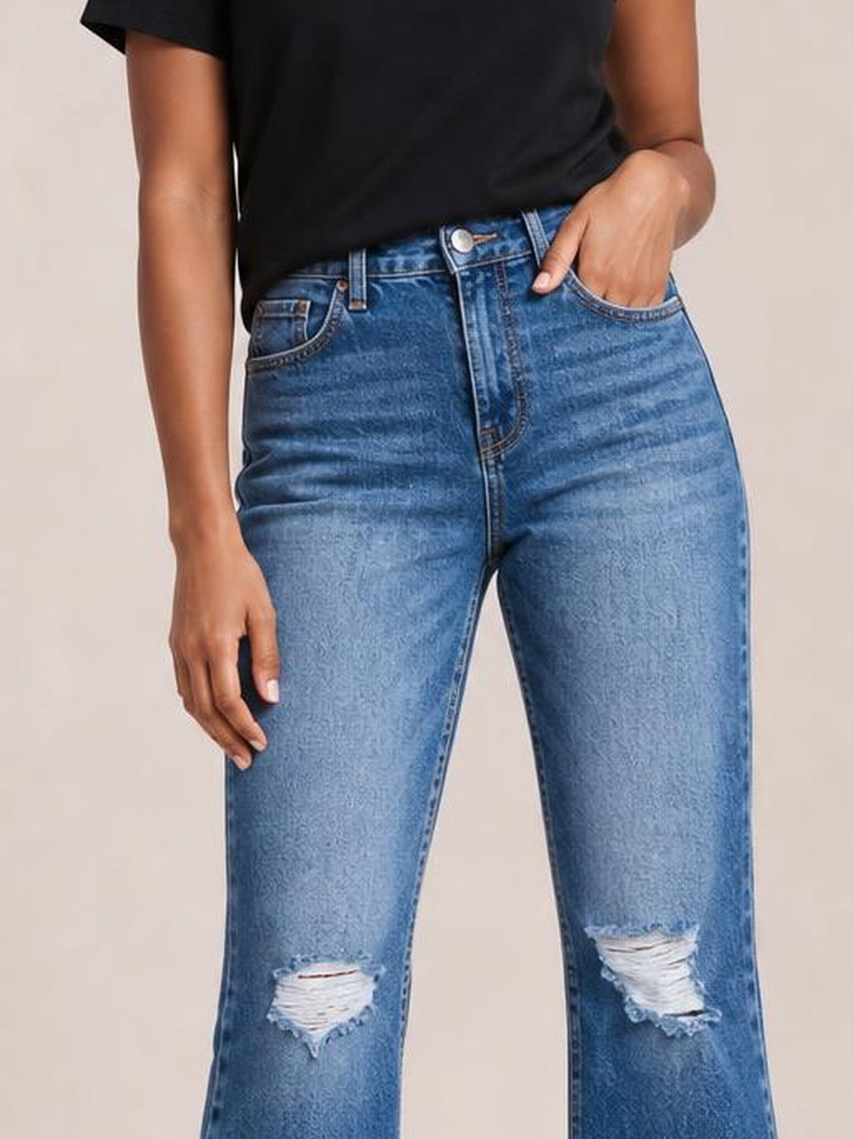A model wears blue high-rise distressed bootcut jeans with a structured waistband, belt loops, front pockets, button fly, and ripped knees.