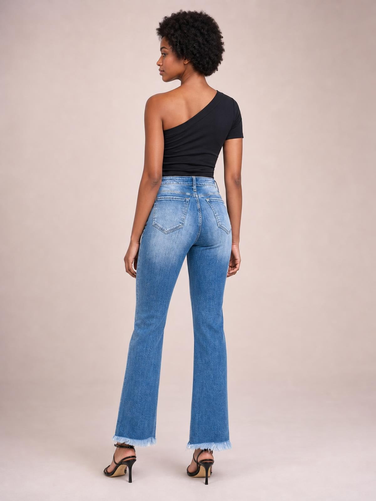 A model poses from behind in light-wash high-rise bootcut jeans with back pockets, a fitted silhouette, subtle distressing, and raw hems.