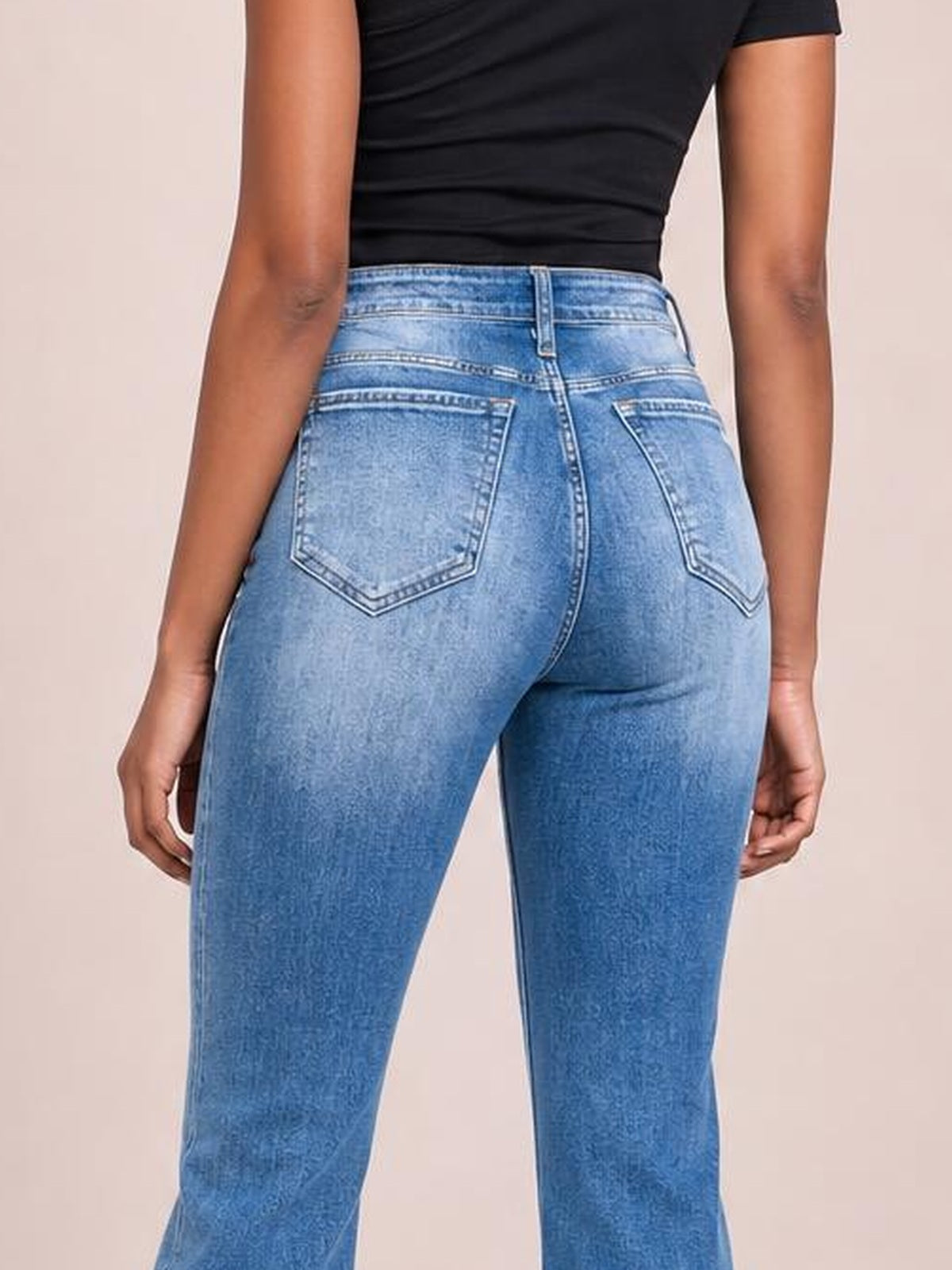 A model stands facing away in light blue high-rise bootcut jeans with a fitted silhouette, belt loops, contrast stitching, and two back patch pockets.
