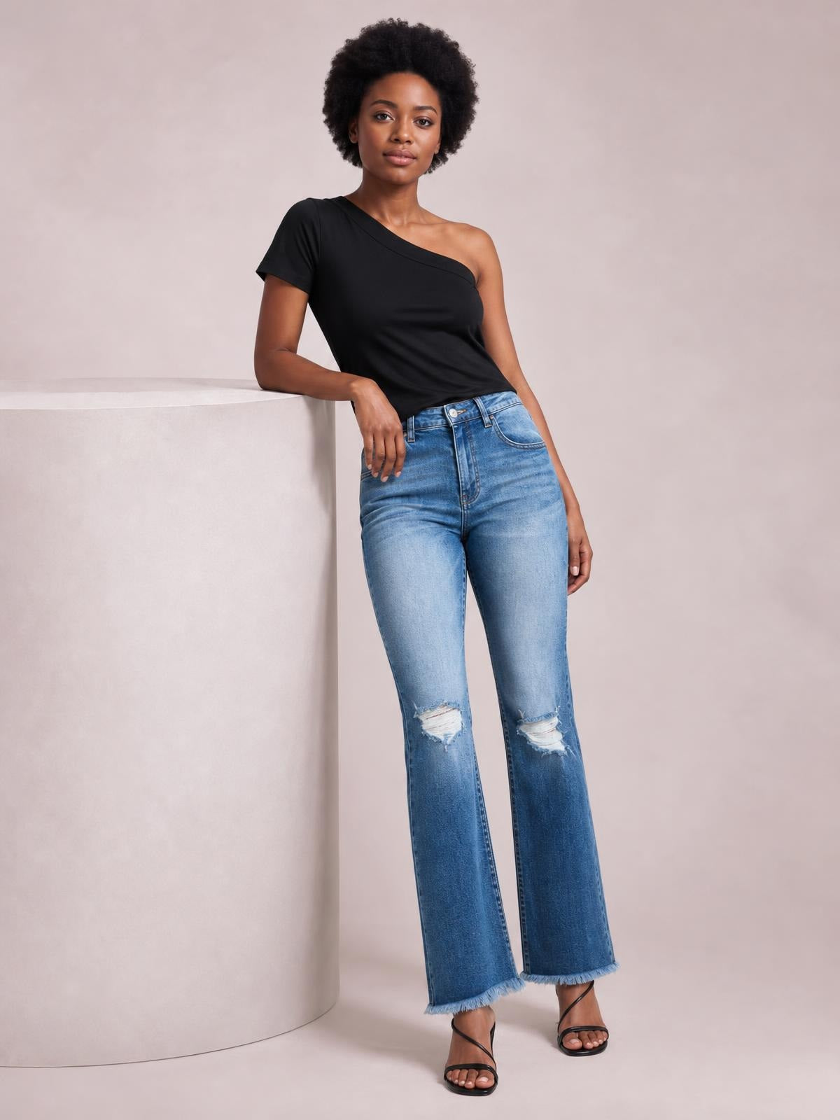 A model leans casually while wearing light-blue high-rise bootcut jeans with five-pocket construction, distressed knee details, and raw frayed hems.