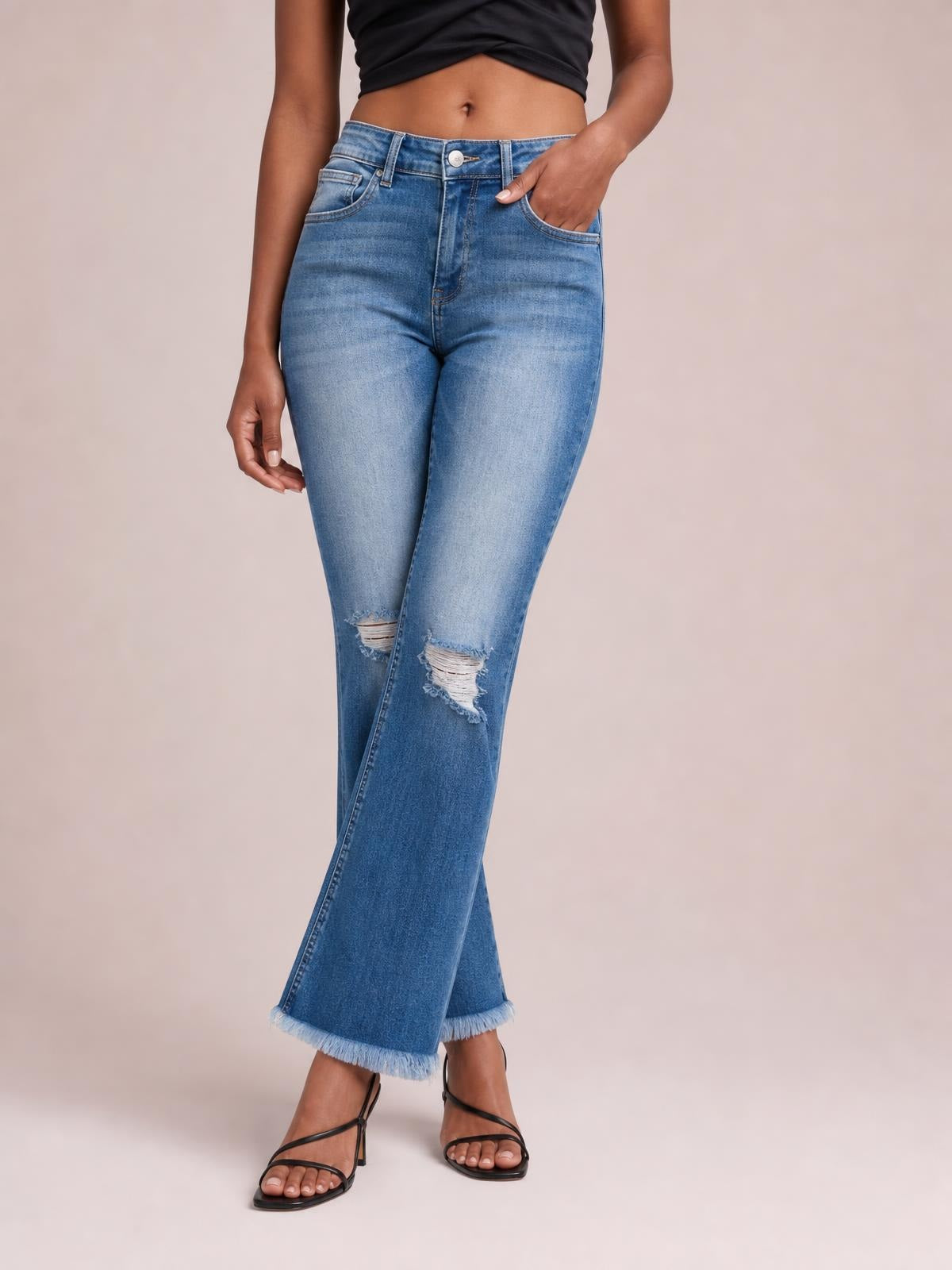 A model poses in light-blue high-rise bootcut jeans with front pockets, distressed knees, and cropped raw hems, one hand tucked into a pocket.