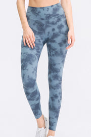 A model wears blue-gray tie-dye slim-fit sports leggings with a wide waistband, long tapered legs, and a curved seam below the waistband while standing with one ankle crossed.