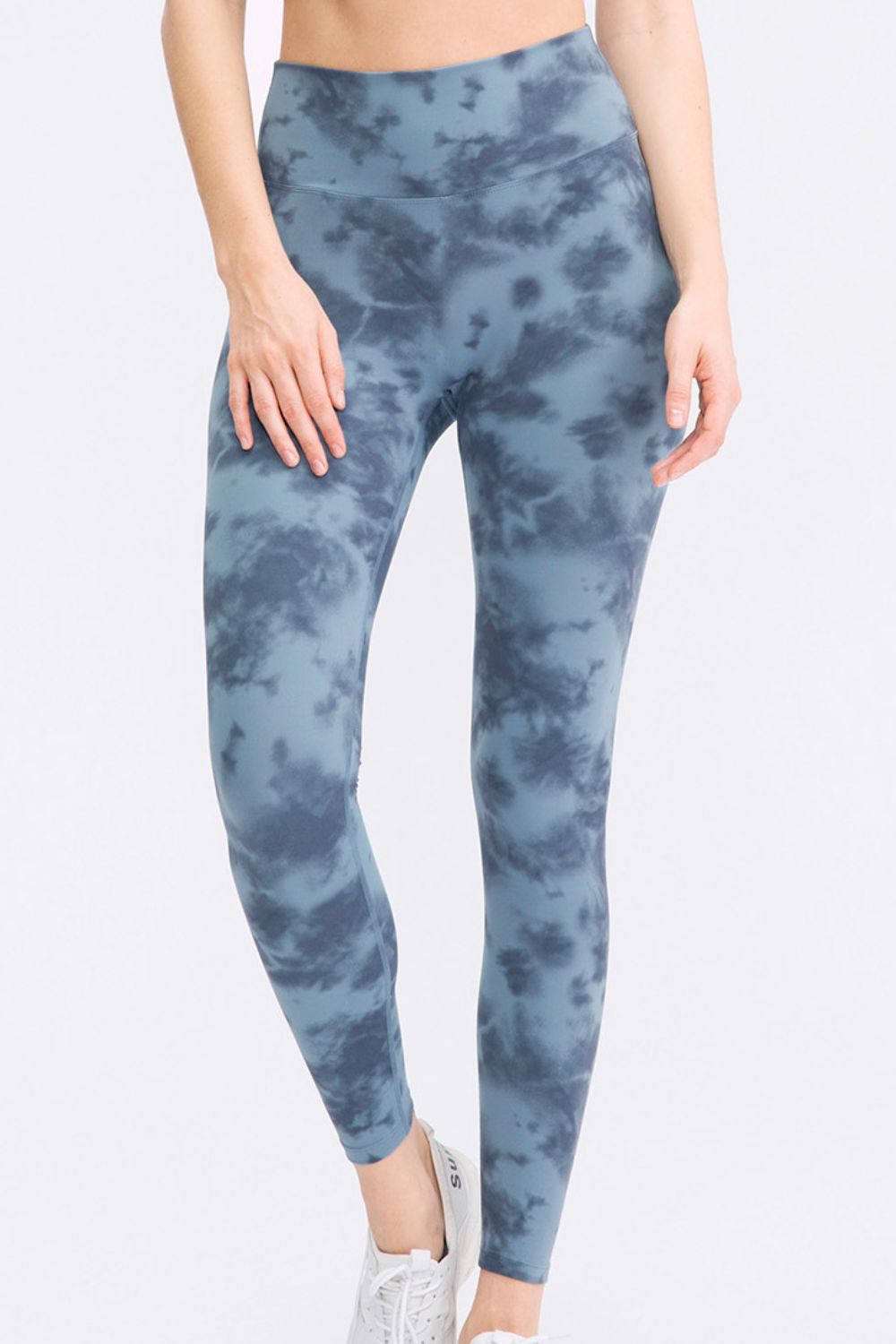 A model wears blue-gray tie-dye slim-fit sports leggings with a wide waistband, long tapered legs, and a curved seam below the waistband while standing with one ankle crossed.