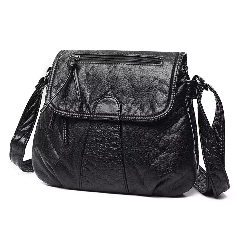 Small black PU leather crossbody bag with an adjustable strap, rounded flap closure, top zip pocket, and stitched panel construction.