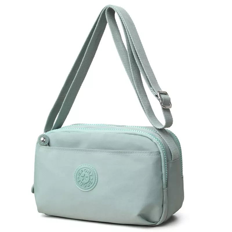 Small mint-green nylon crossbody bag with an adjustable strap, zippered top, front slip pocket, rounded corners, and compact structured silhouette.