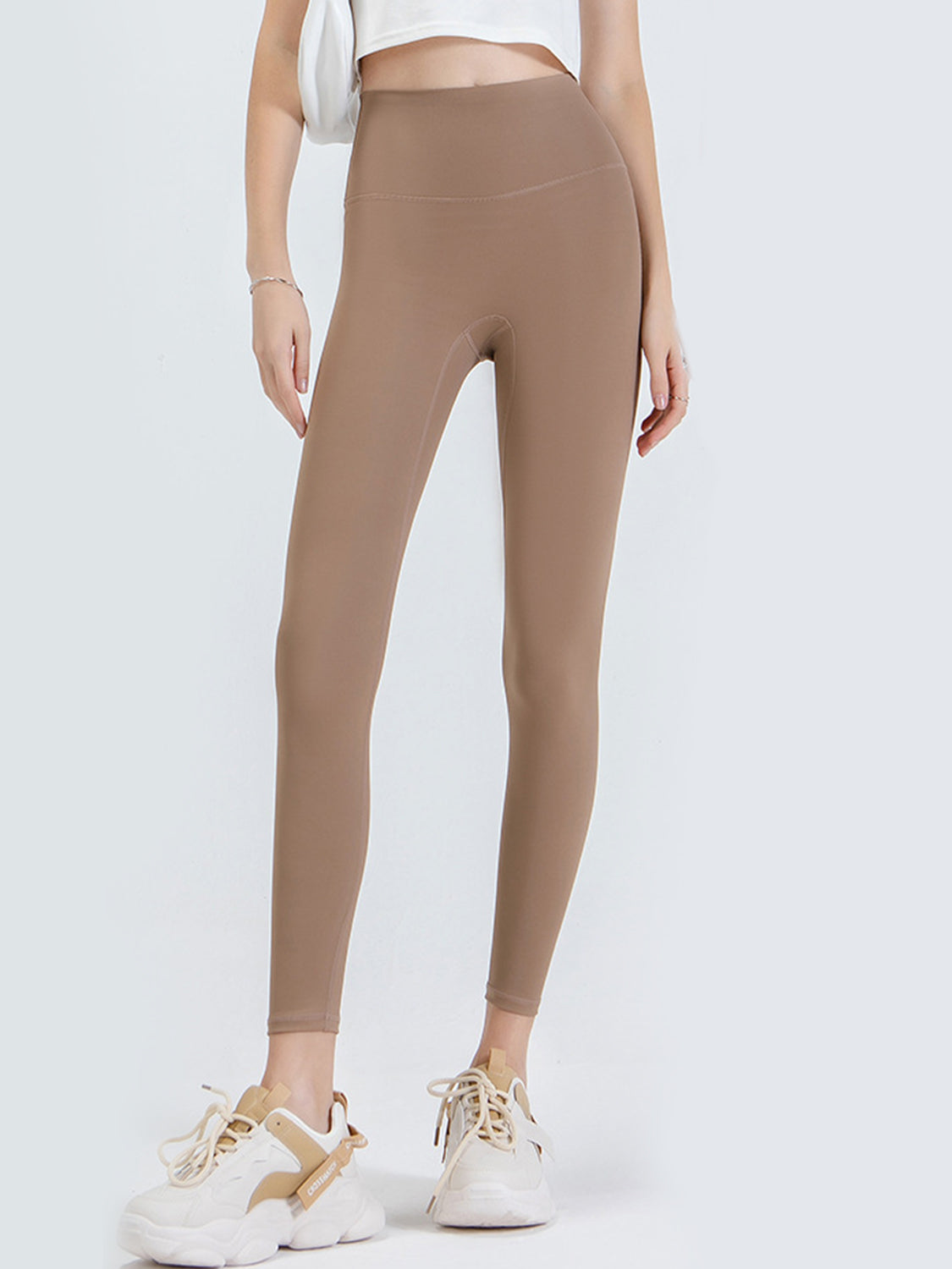 A model stands in taupe sports leggings with a wide high-rise waistband, smooth fitted silhouette, full-length legs, and visible curved seam construction.