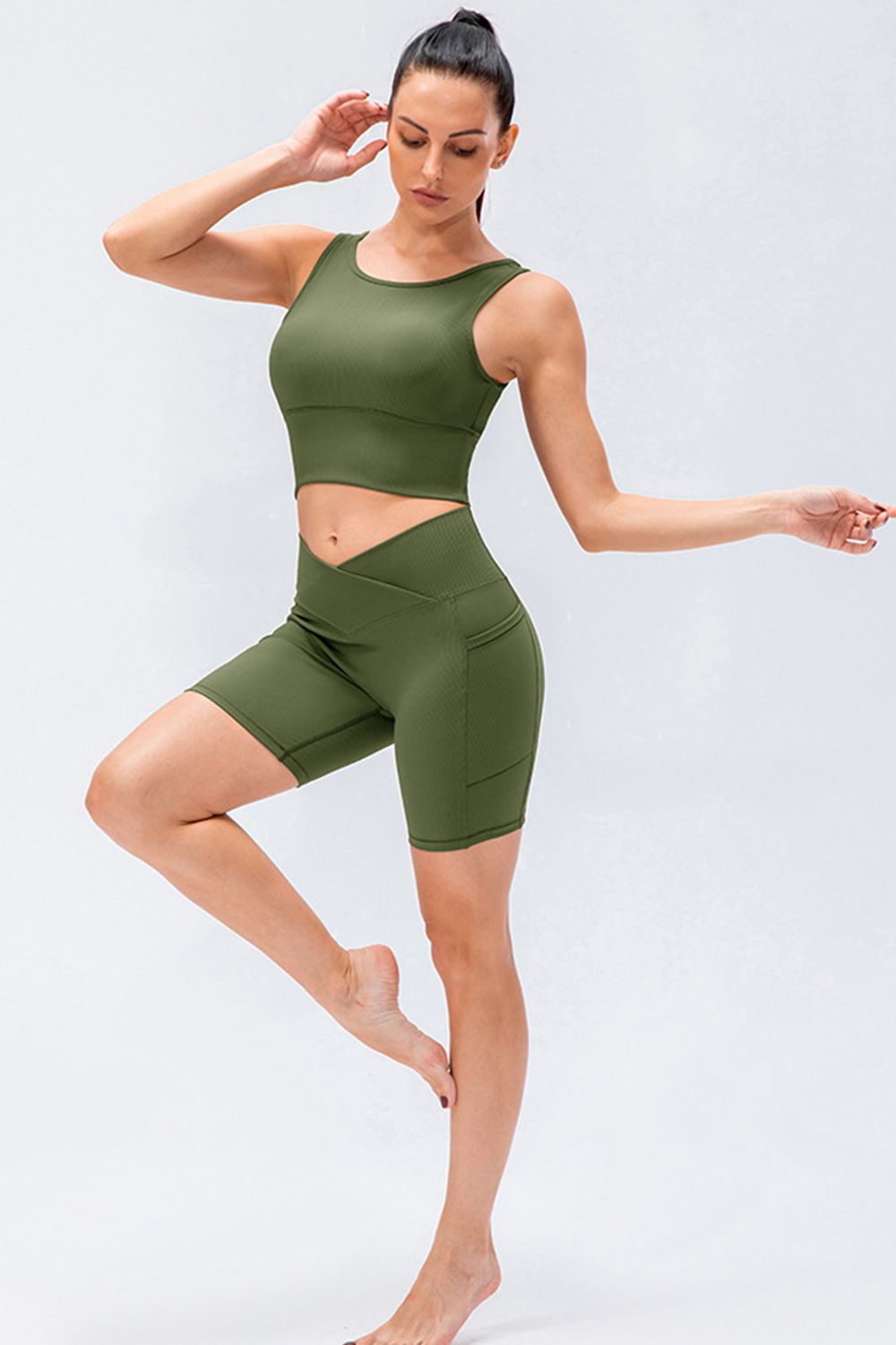 Model poses on one leg wearing solid olive-green slim-fit sports shorts with a crossover V-waistband, stretchy fitted silhouette, and knee-length construction.