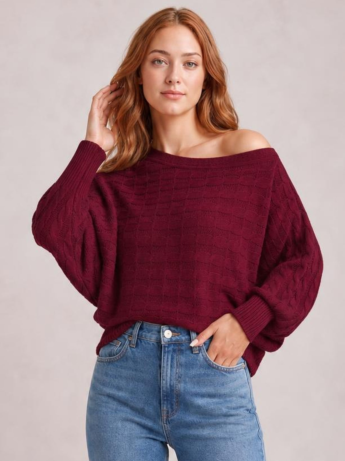 Model wearing a burgundy cable-knit boat neck sweater with an oversized silhouette, dropped shoulders, voluminous sleeves, and ribbed cuffs, posing with one hand in her jeans pocket.