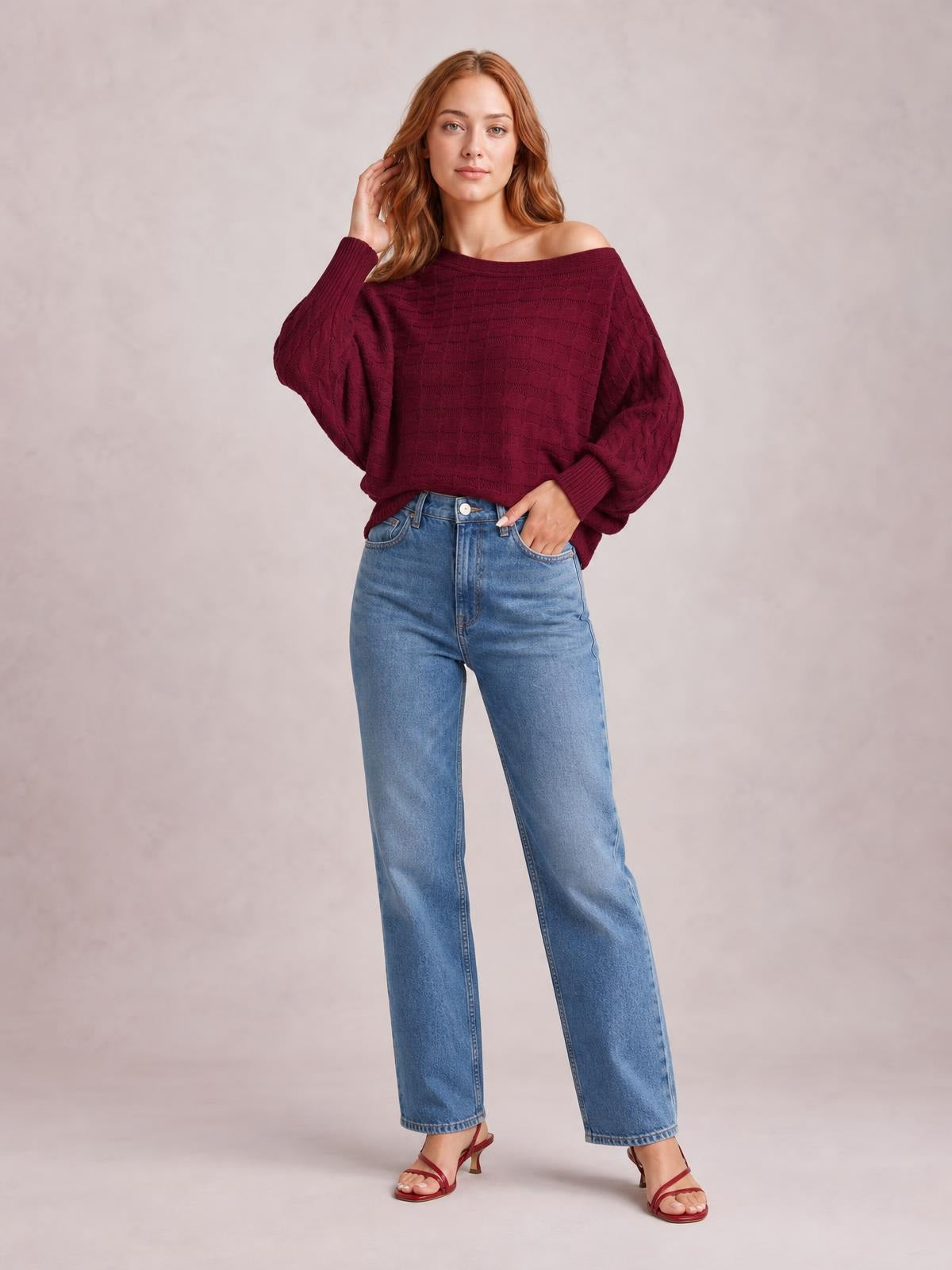 A model poses in a burgundy cable-knit boat neck sweater with a relaxed oversized silhouette, long sleeves, ribbed cuffs, and a wide neckline.
