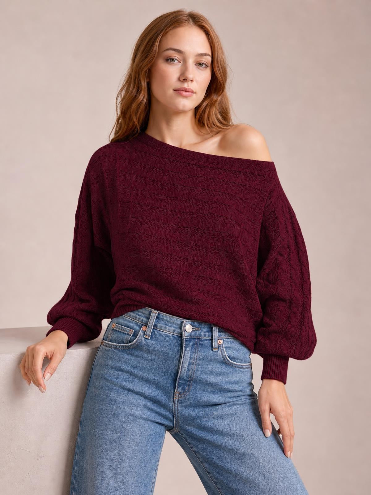 Model poses in a burgundy cable-knit boat neck sweater with an off-shoulder neckline, relaxed silhouette, balloon sleeves, and ribbed cuffs.