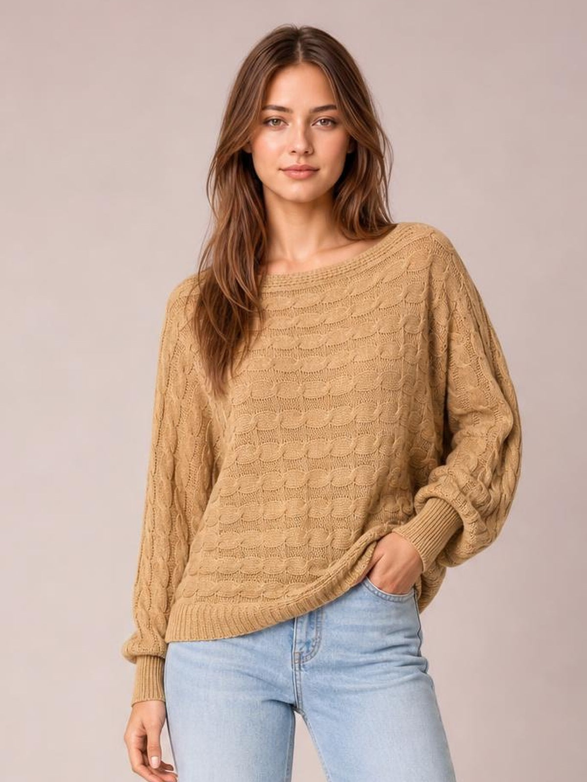 Model wears a camel cable-knit boat neck sweater with a relaxed silhouette, long sleeves, ribbed cuffs and hem, posing with one hand in her jeans pocket.
