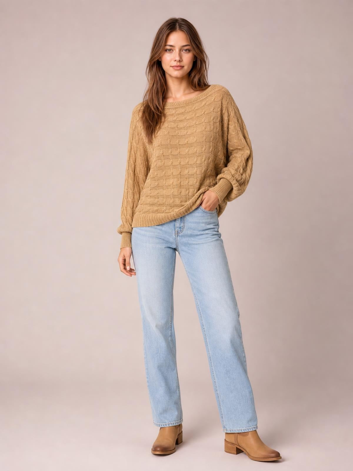 A model wears a camel cable-knit boat neck sweater with a relaxed silhouette, long sleeves, ribbed cuffs and hem, posing with one hand in her jeans pocket.