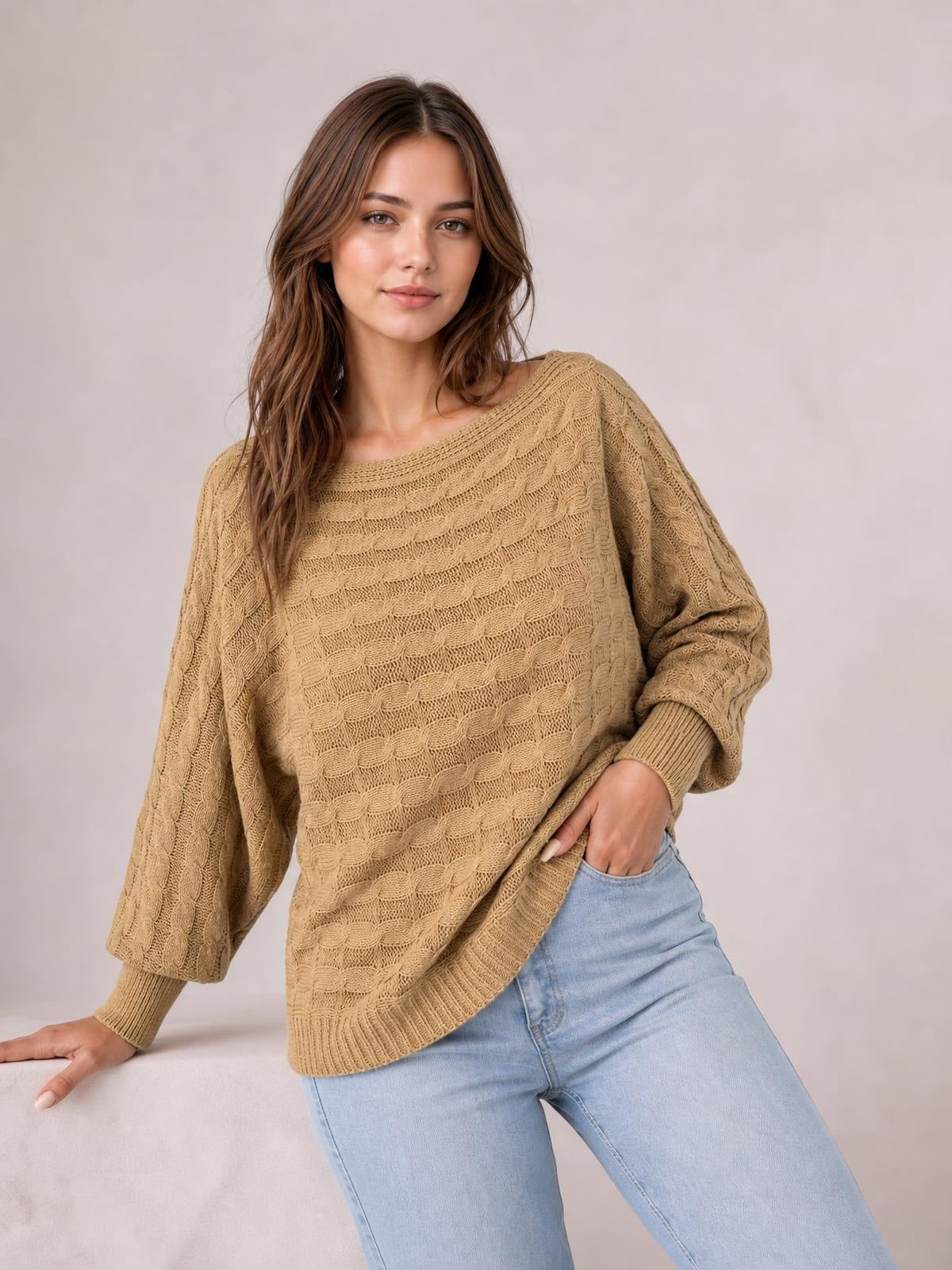 Model poses in a relaxed stance wearing a tan cable-knit boat neck sweater with a loose silhouette, ribbed cuffs and hem, and balloon sleeves.