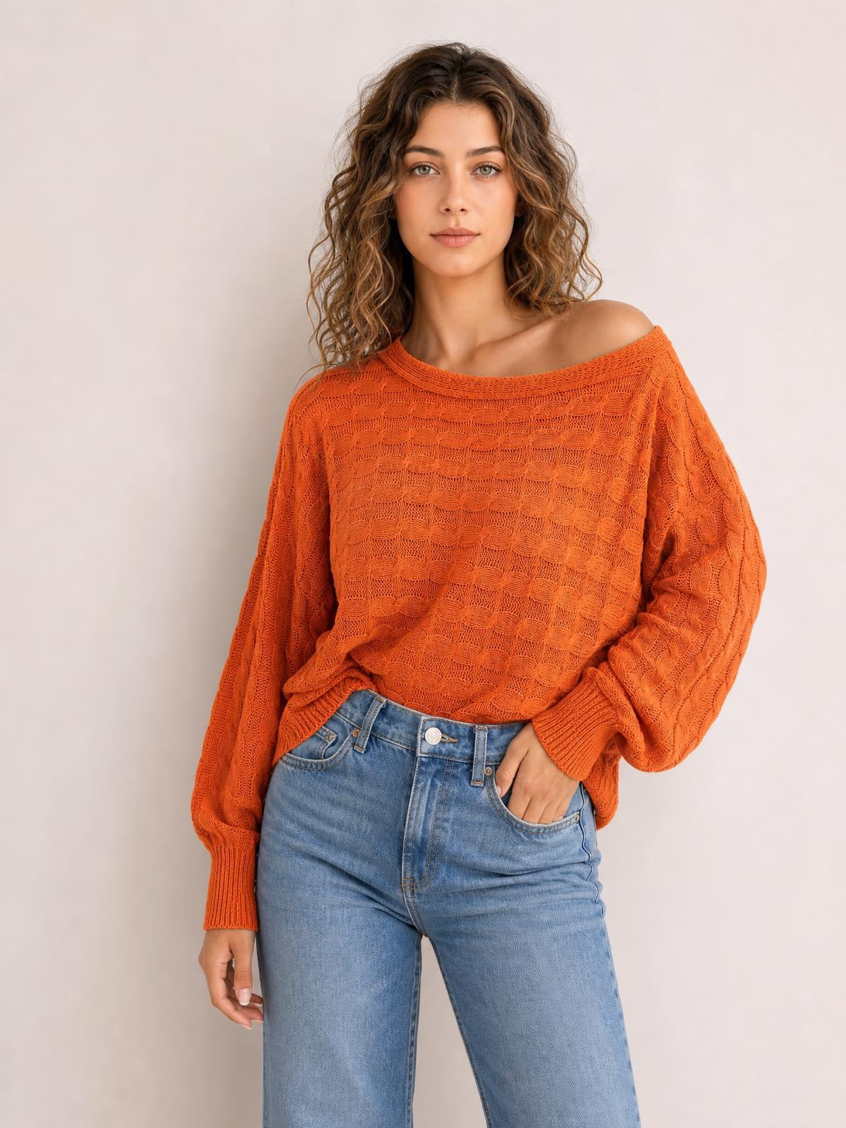 Model wears an orange cable-knit boat neck sweater with a relaxed oversized silhouette, long sleeves, ribbed cuffs, and one hand tucked into her jeans pocket.