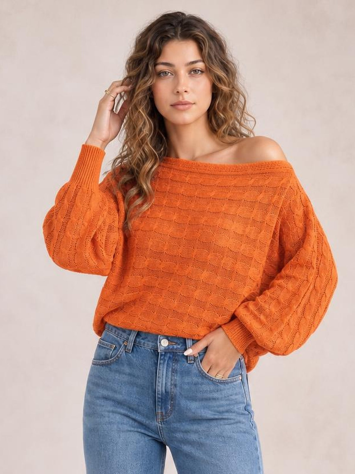 Model wearing an orange cable-knit boat neck sweater with an off-shoulder neckline, relaxed silhouette, and voluminous sleeves, posing with one hand in her jeans pocket.