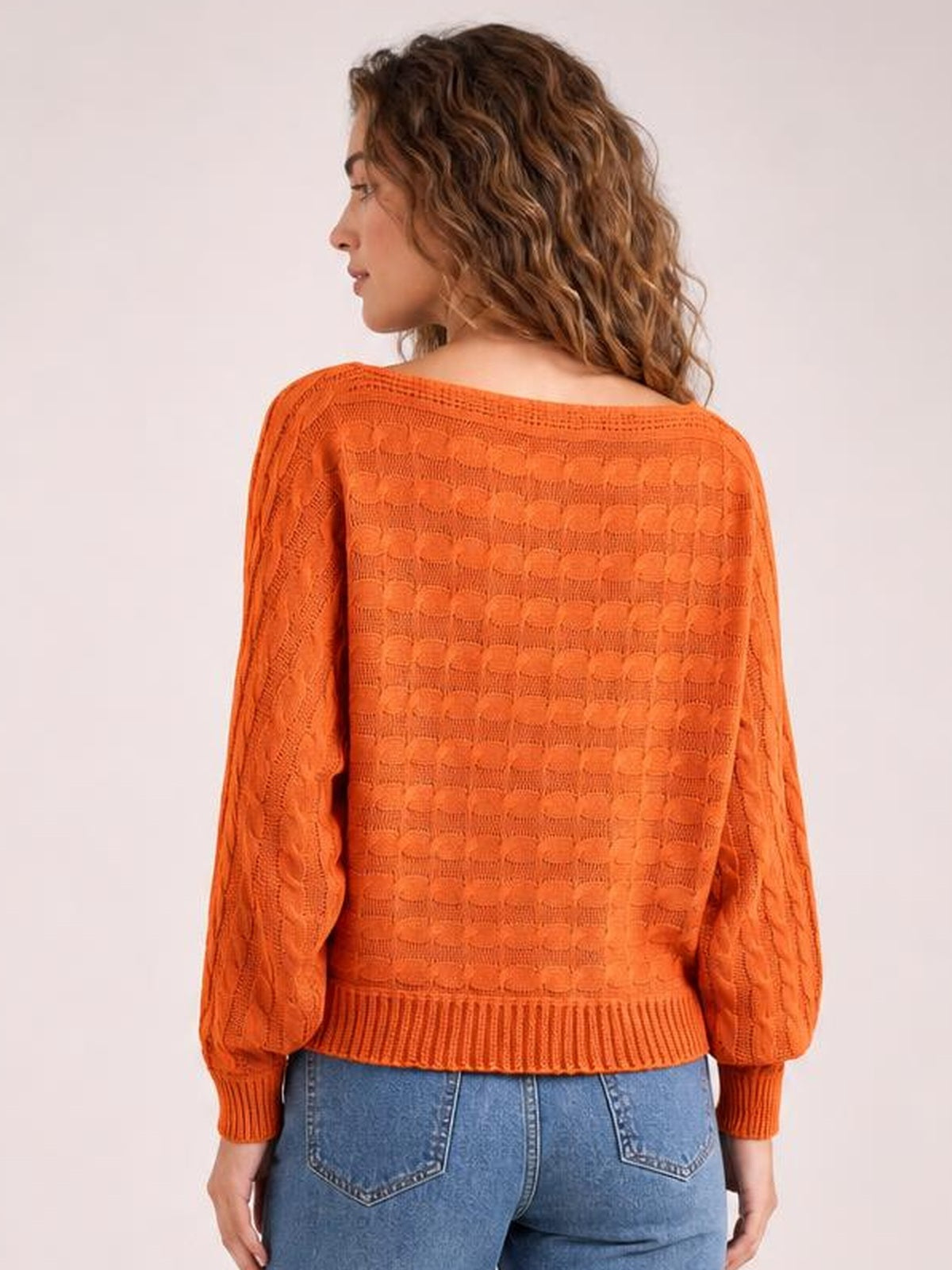 Model shown from behind wears an orange cable-knit boat neck sweater with a relaxed silhouette, long sleeves, ribbed cuffs, and a ribbed hem.